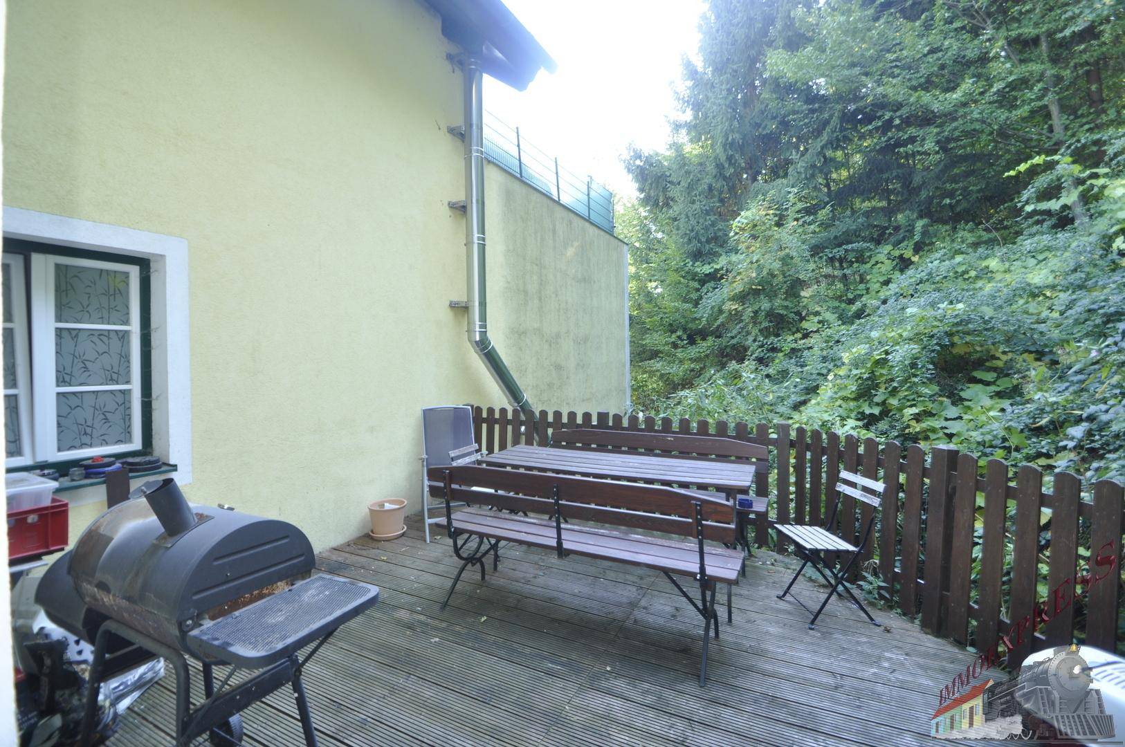 Holzterrasse mit Grillbereich, Sitzgelegenheiten und umgeben von viel Grün.