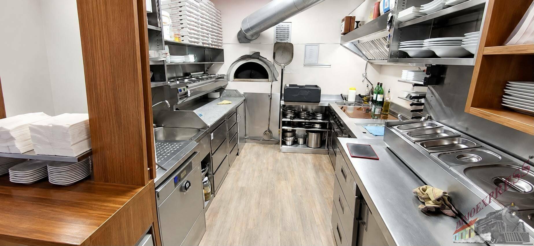 Geräumige Industrieküche mit professionellen Edelstahlgeräten, Pizzaofen und Holzregalen.