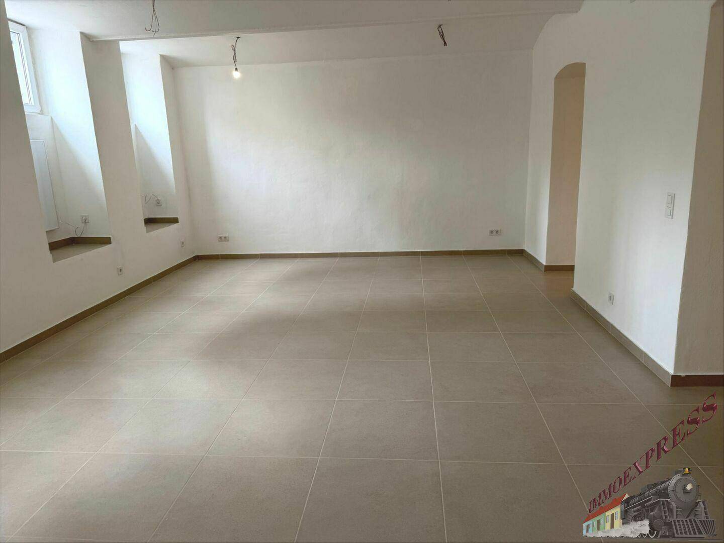 Heller, leerer Raum mit Fliesenboden und einem Bogen, der zu einem weiteren Bereich führt.