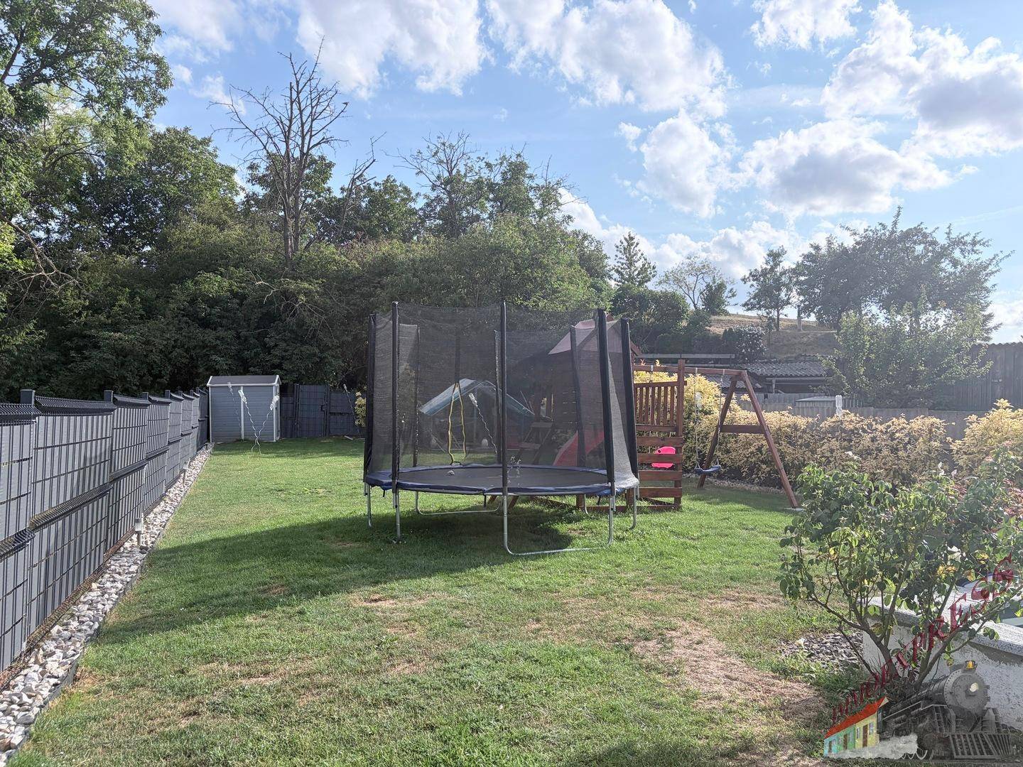 Hinterer Gartenbereich mit Trampolin, Schaukel und Gartenhaus umgeben von Bäumen.