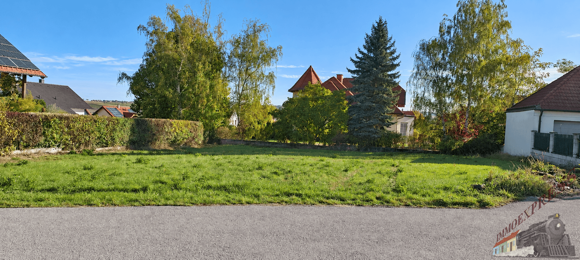 Blick von der Straße auf ein unbebautes Grundstück mit grüner Wiese und umgebender Bebauung.