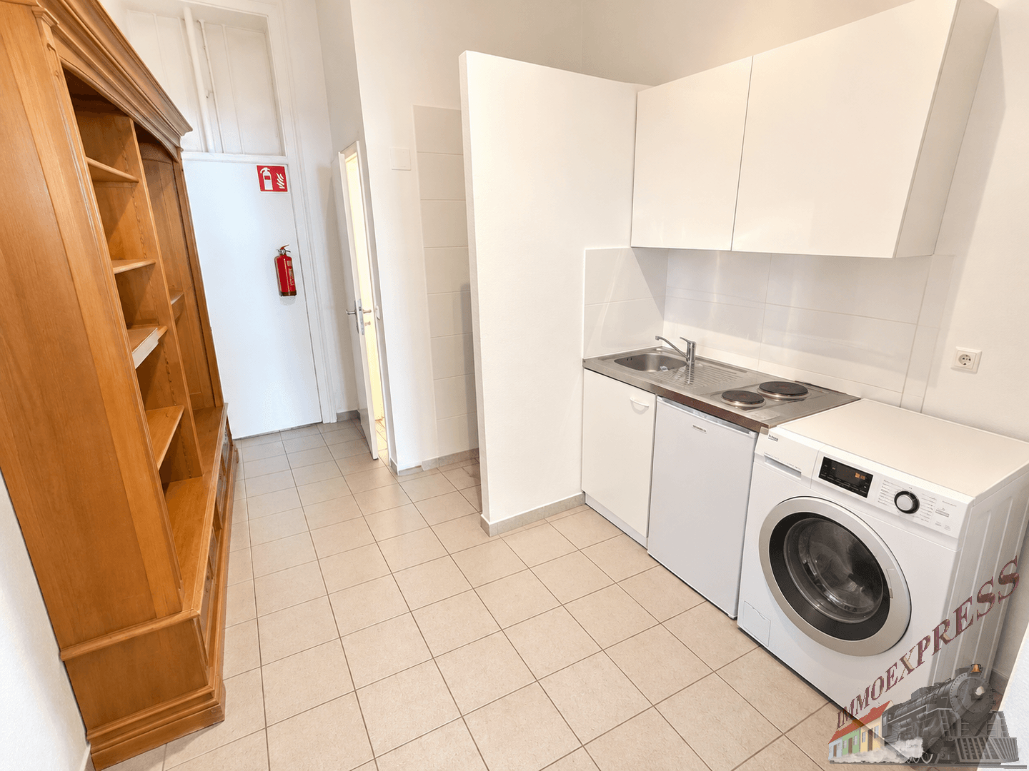 Kompakte Küchenzeile mit Waschmaschine, Spüle und Kochplatten, ergänzt durch ein großes Holzregal.