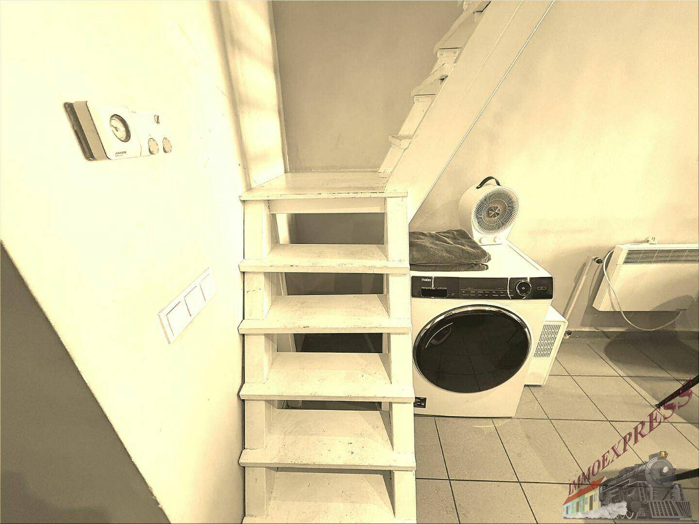 Bereich unter der Treppe mit Waschmaschine, Trockner und Heizung auf gefliestem Boden.
