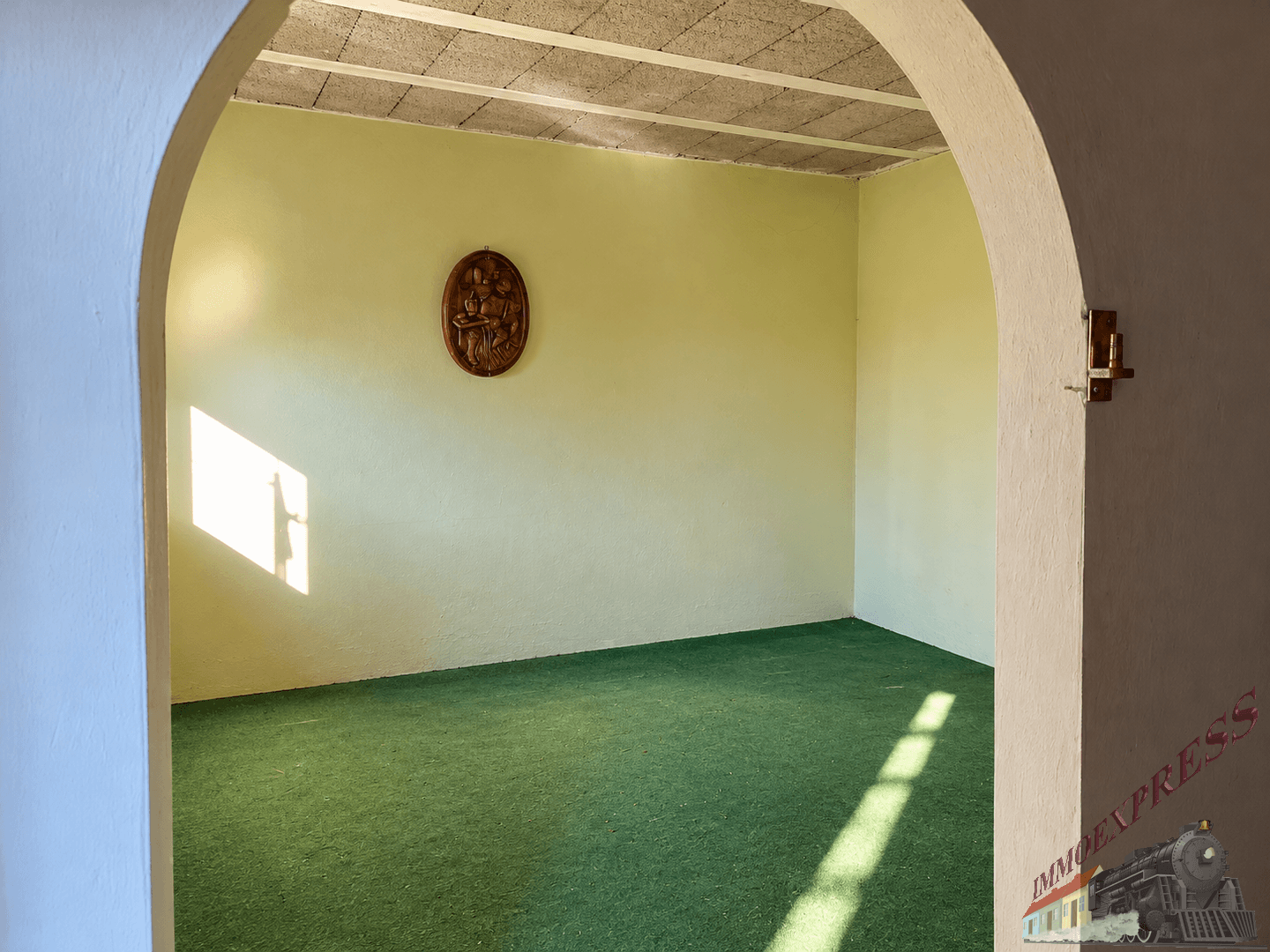 Heller Innenraum mit grünem Teppichboden und einem Rundbogen, durch den Sonnenlicht fällt.