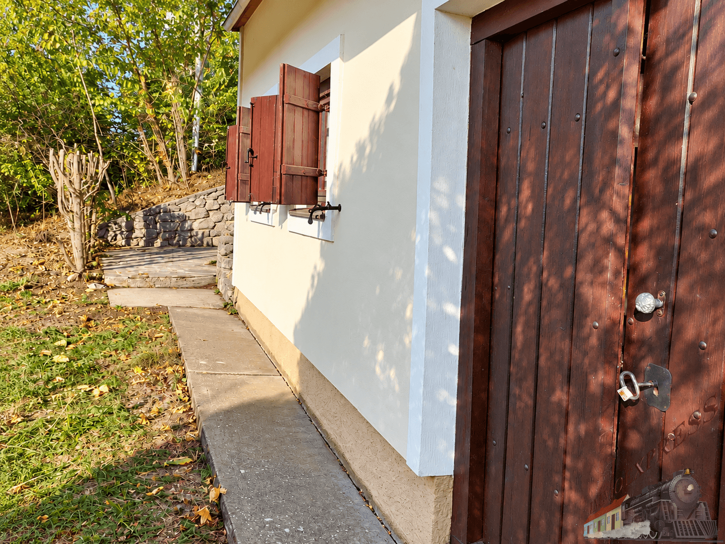 Detailansicht der hellen Fassade mit rustikaler Holztür und geöffneten Fensterläden, umgeben von Bäumen.