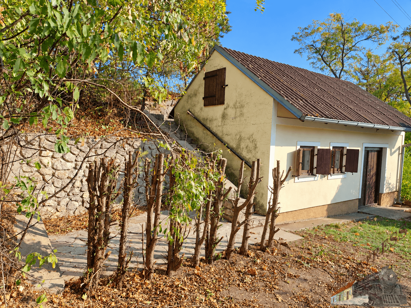 Seitenansicht eines kleinen Gebäudes mit Steintreppe, umgeben von herbstlicher Vegetation und Bäumen.