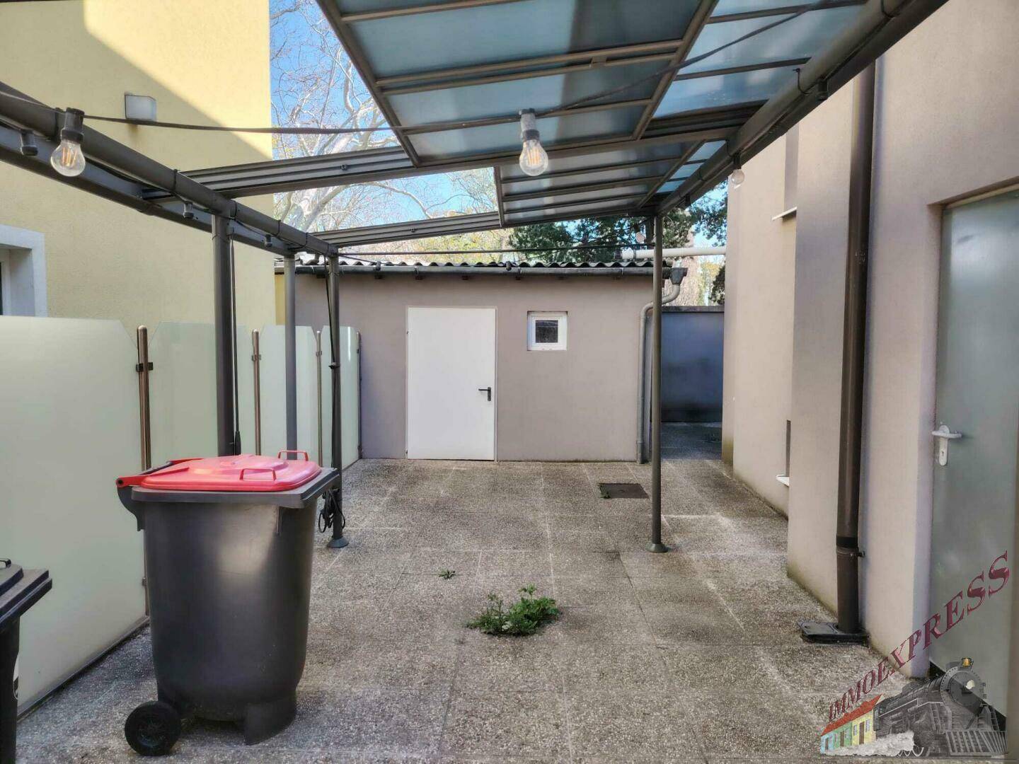 Überdachter Außenbereich mit Betonpflaster und einem kleinen Schuppen im Hintergrund.