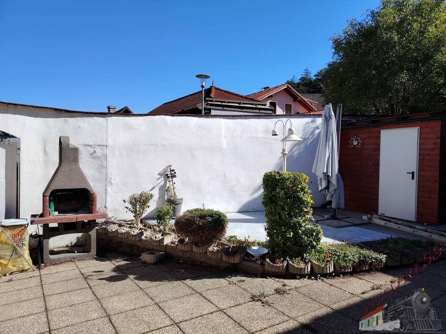 Geräumige gepflasterte Terrasse mit festem Grill, Gartenhaus und einer hohen weißen Mauer. Ideal für Feste.