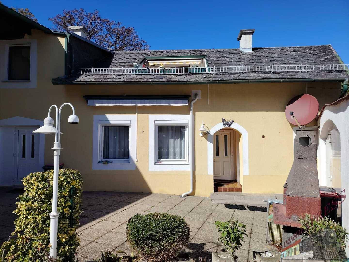 Sonniger Terrassenbereich vor dem gelben Haus mit Grill und dekorativer Außenbeleuchtung. Einladender Eingangsbereich.
