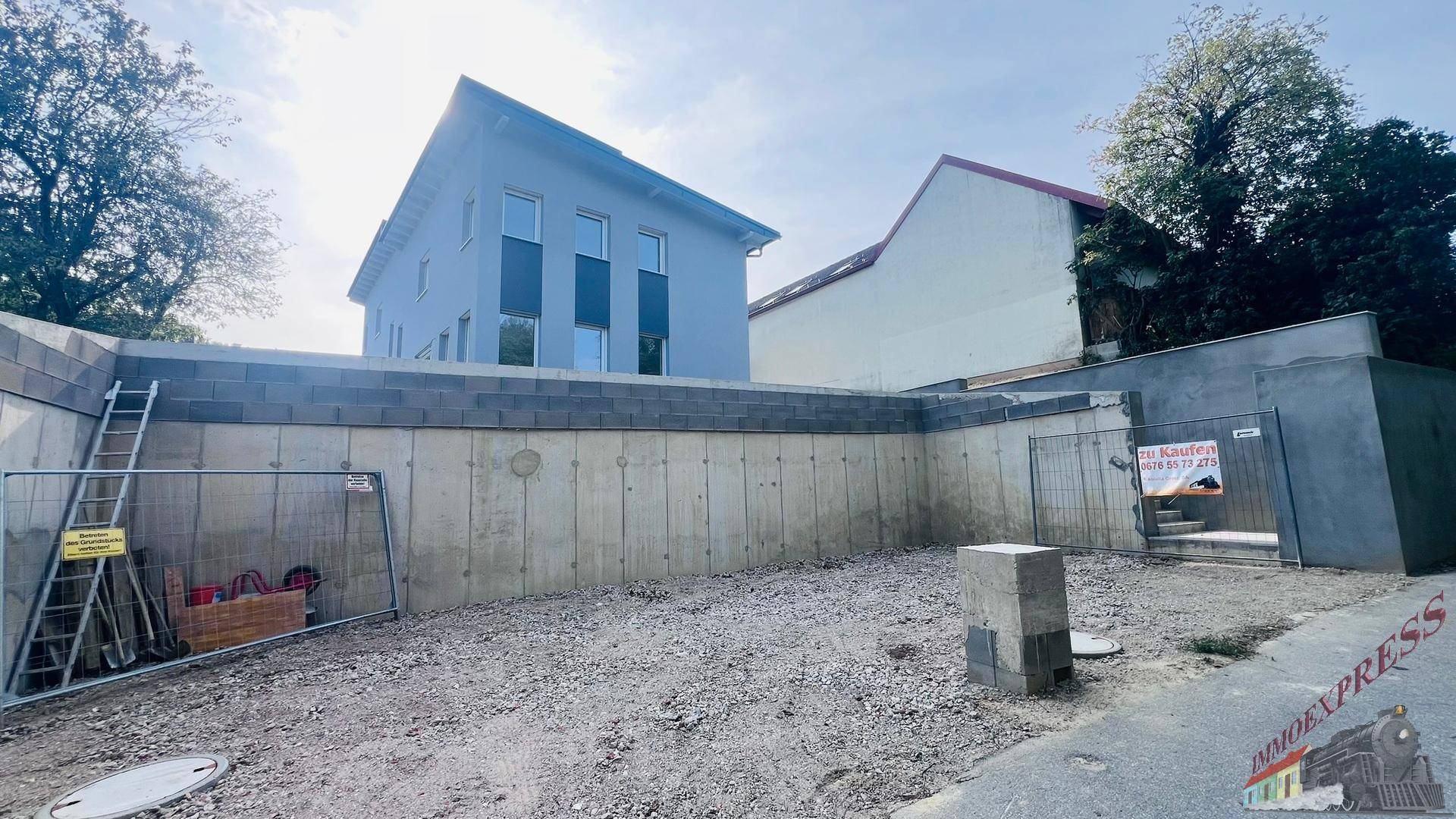 Blick auf die Baustelle mit einer hohen Betonmauer und dem Neubau im Hintergrund.