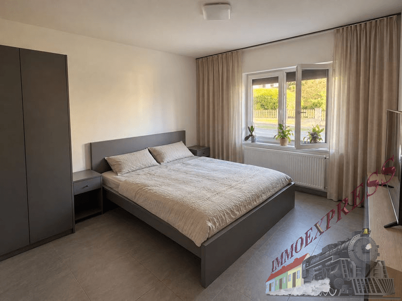 Helles Schlafzimmer mit grauem Doppelbett, passendem Kleiderschrank und Fliesenboden.