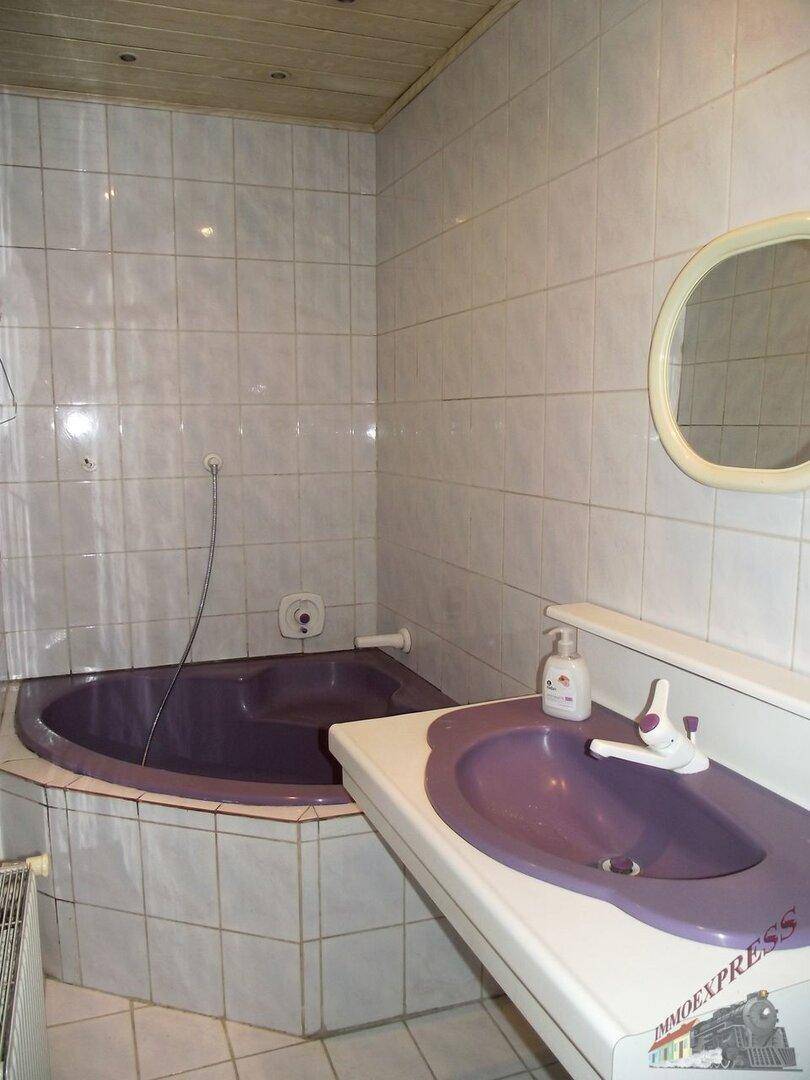 Badezimmer im 80er-Jahre-Stil mit lila Eckbadewanne und passendem Waschbecken, geflieste Wände.