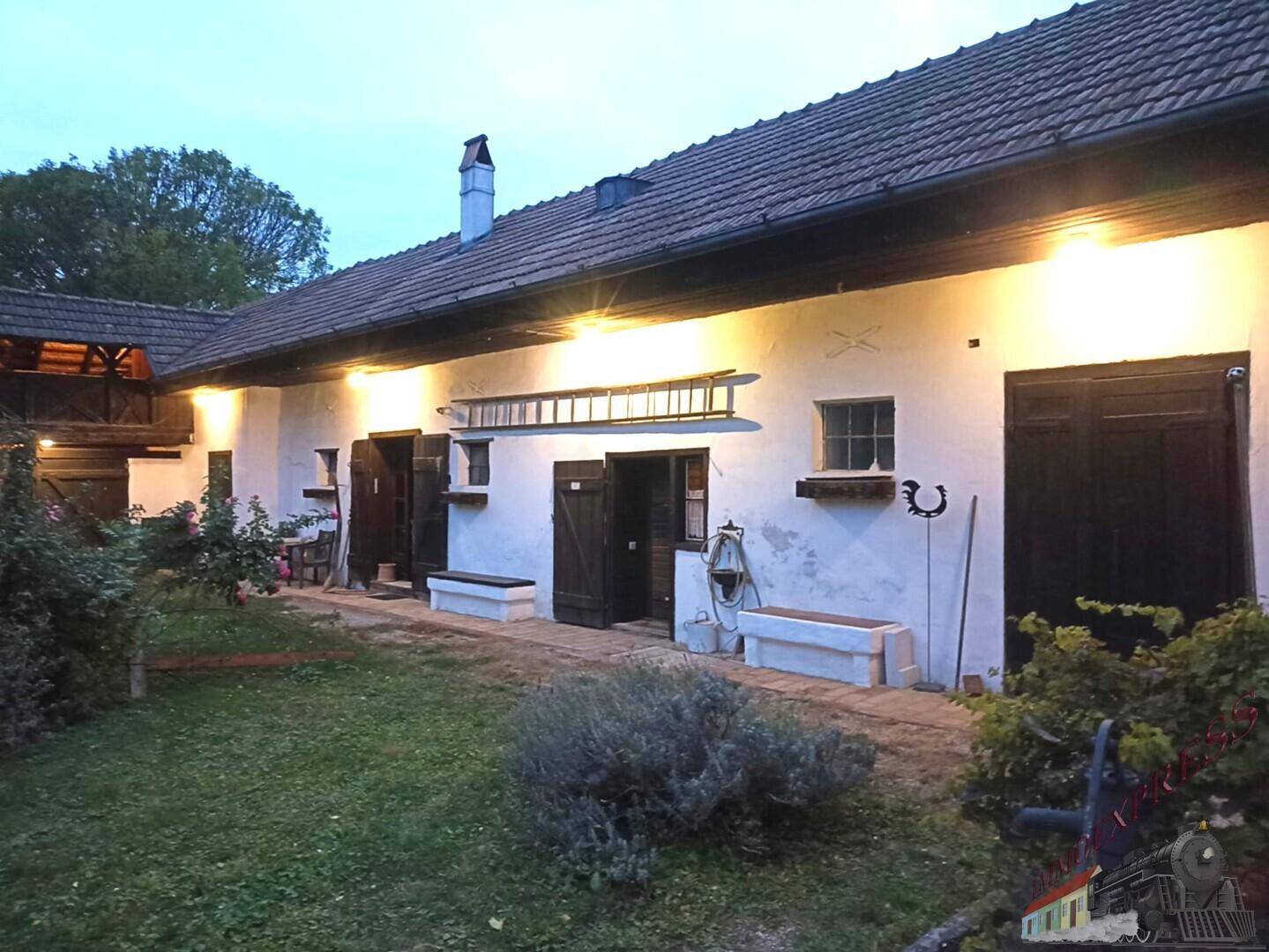 Beleuchteter Gartenbereich am Abend mit ländlichem Haus und Holzfensterläden.