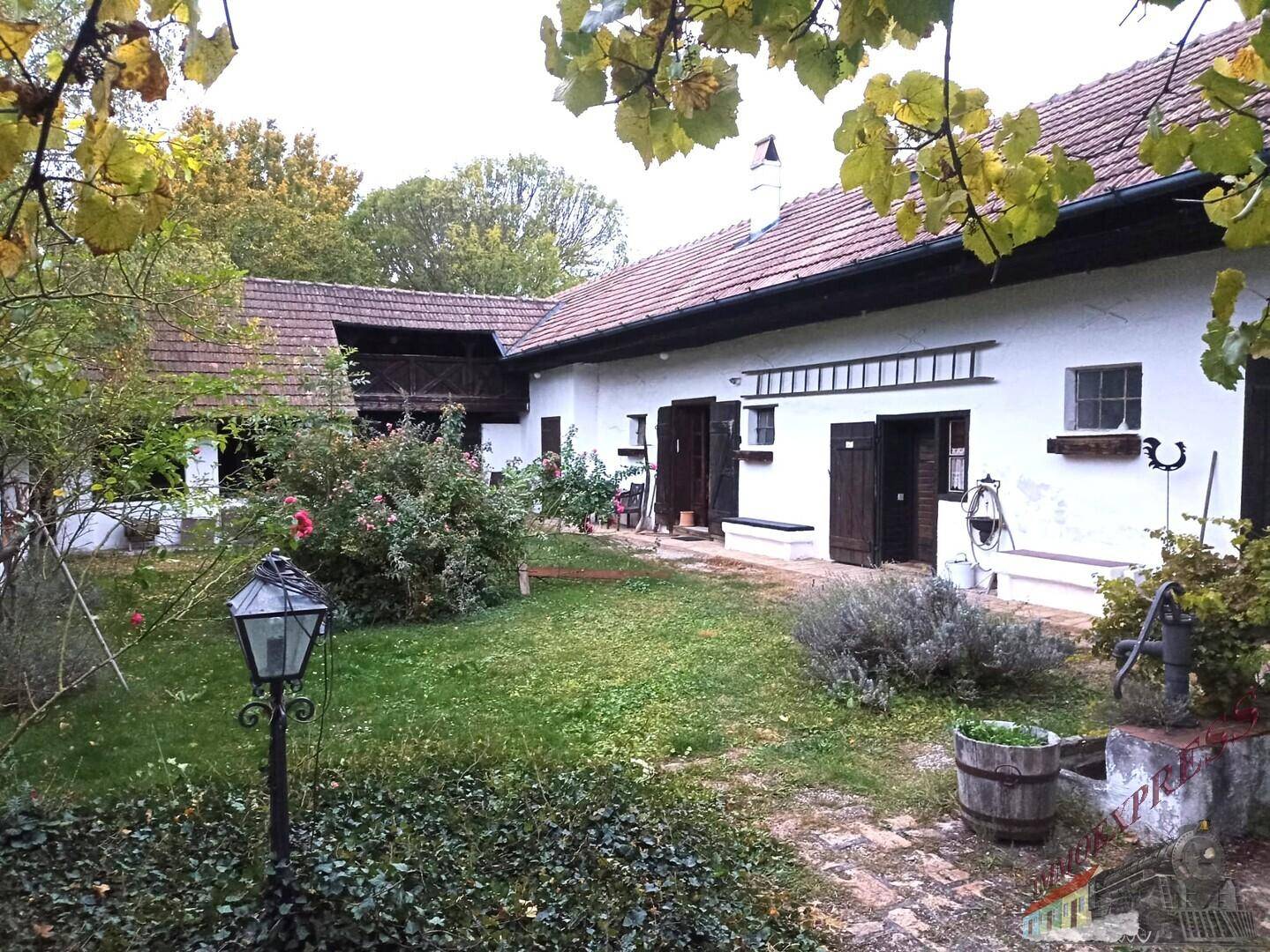 Weitläufiger Gartenbereich mit alten Bäumen, Laterne und ländlichem Haus im Hintergrund.