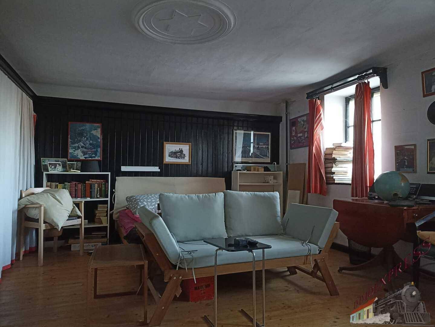 Helles Zimmer mit Schlafsofa, Bücherregal und Schreibtisch am Fenster.