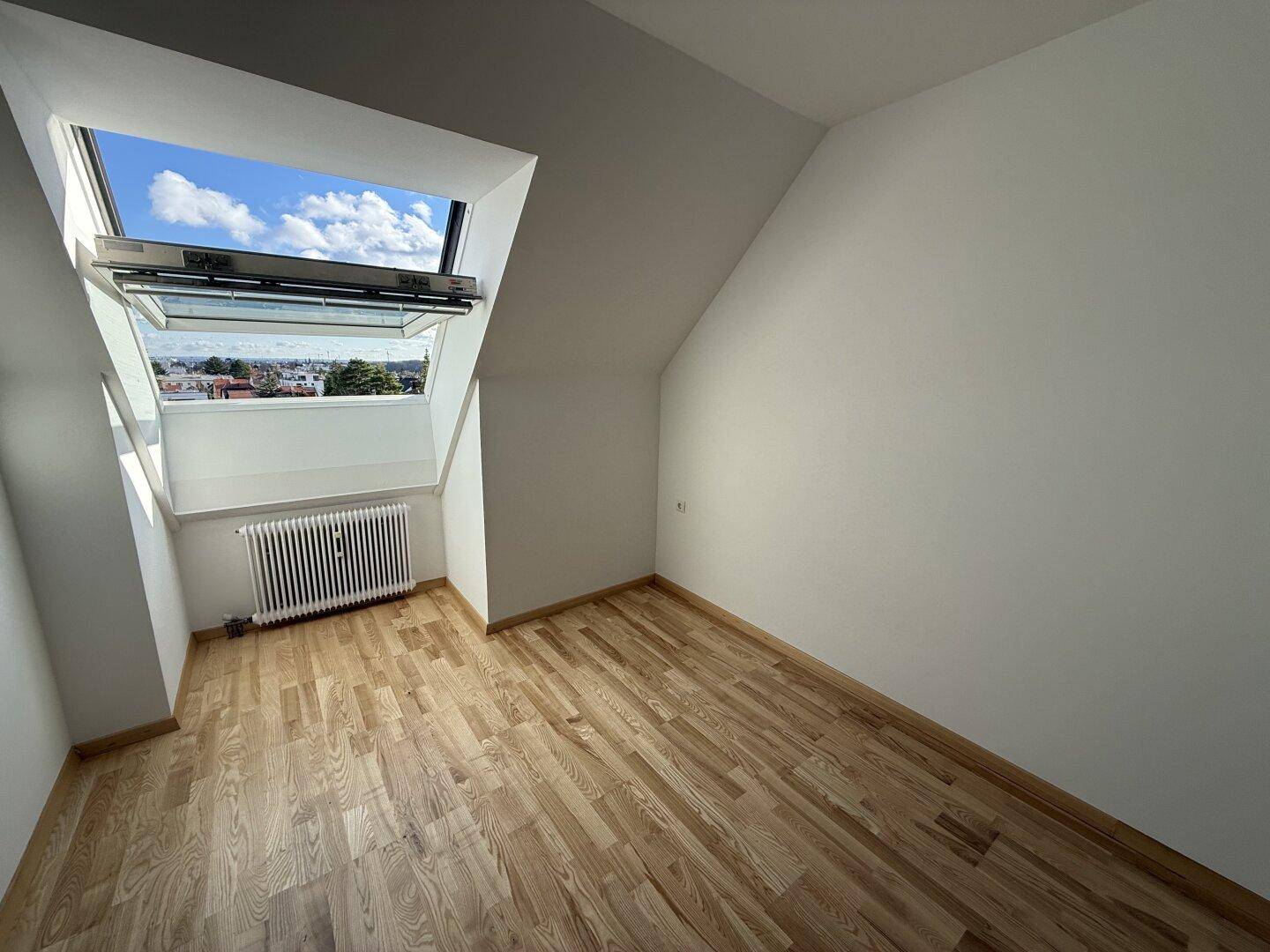 Leerer Raum im Dachgeschoss mit Holzboden, Dachfenster und Heizkörper unter der Schräge.