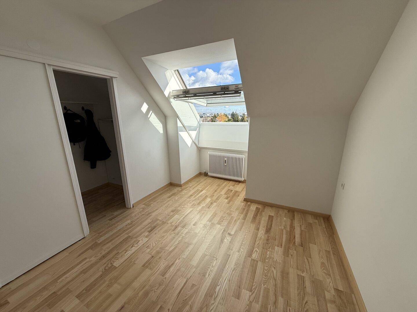 Leeres Dachgeschosszimmer mit Holzboden, Dachfenster und Zugang zu einem begehbaren Kleiderschrank.