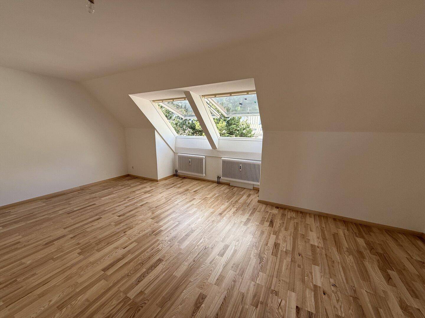 Geräumiges, leeres Wohnzimmer im Dachgeschoss mit Holzboden und zwei großen Dachfenstern.