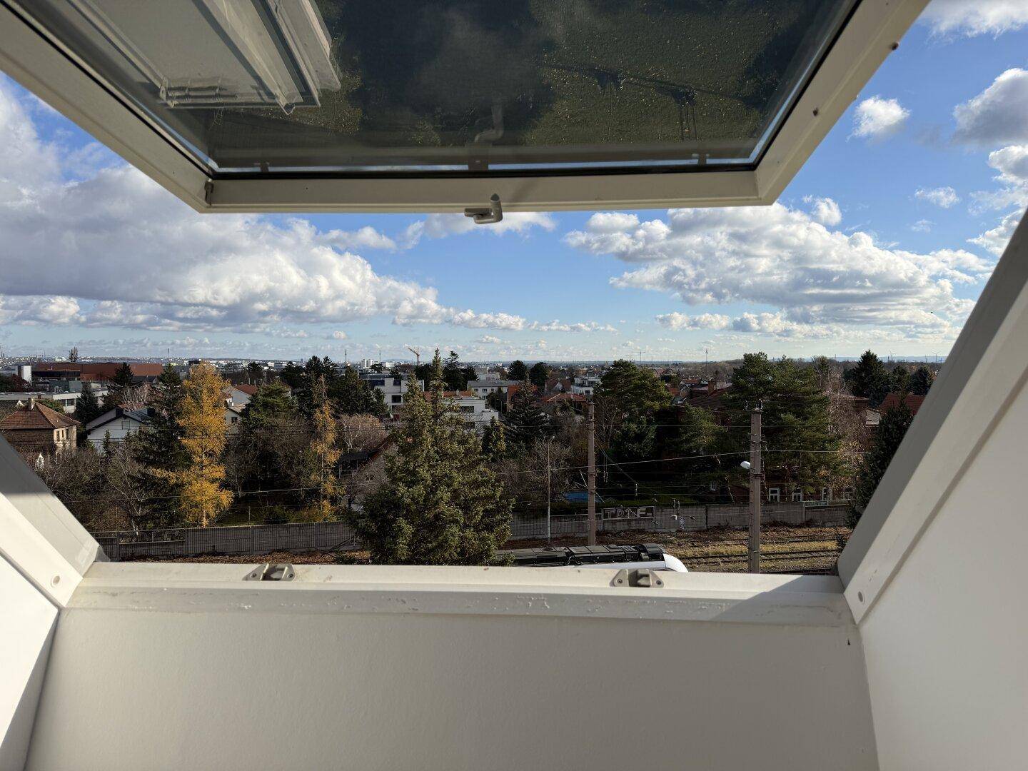 Weitblick über die Stadt und grüne Umgebung durch ein geöffnetes Dachfenster.