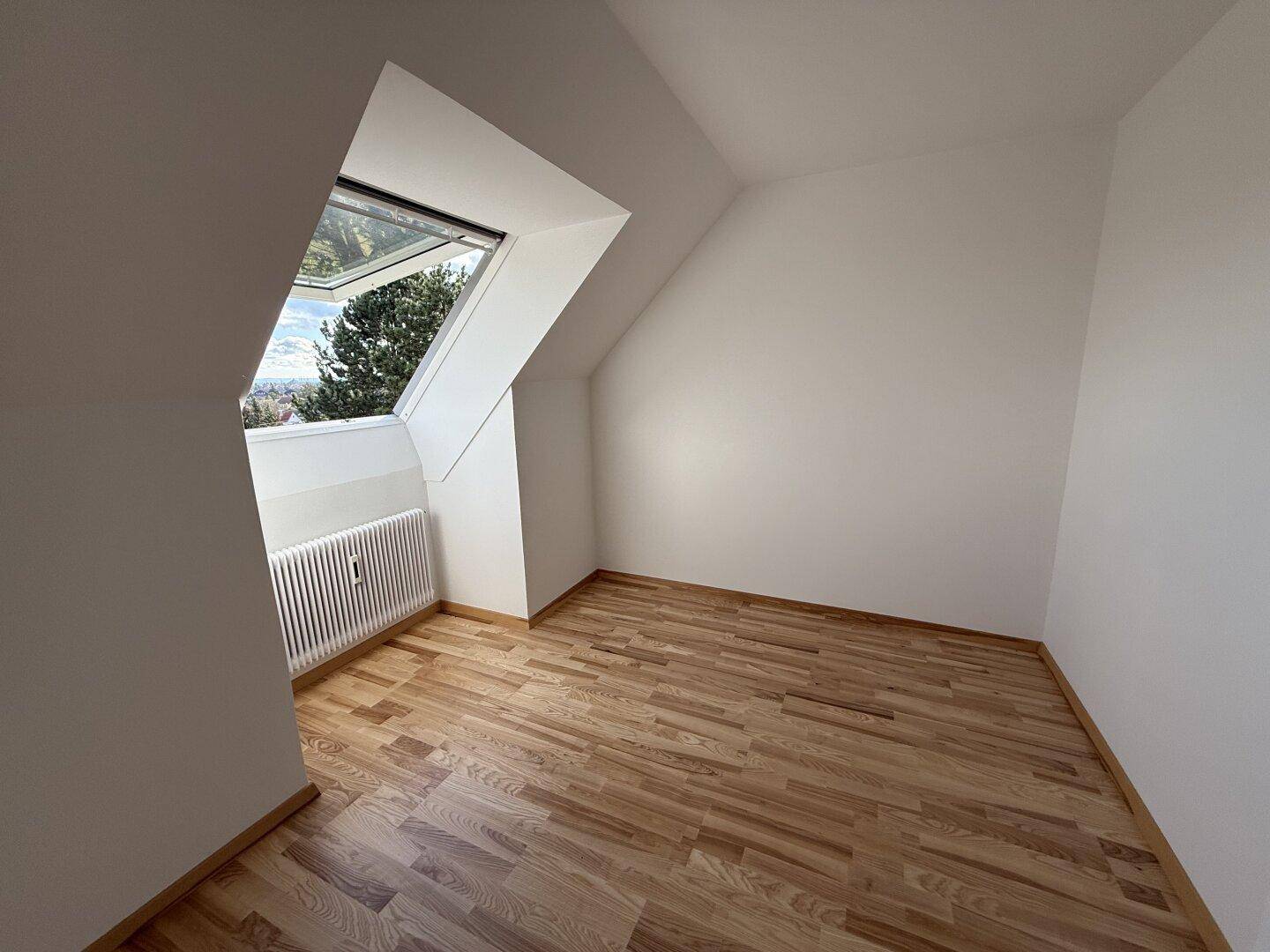 Leeres Schlafzimmer im Dachgeschoss mit Holzboden, Dachfenster und Heizkörper unter der Schräge.