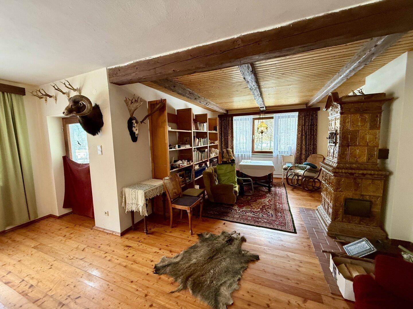 Wohnzimmer mit Holzdielenboden, Kachelofen und rustikalen Holzbalken an der Decke.