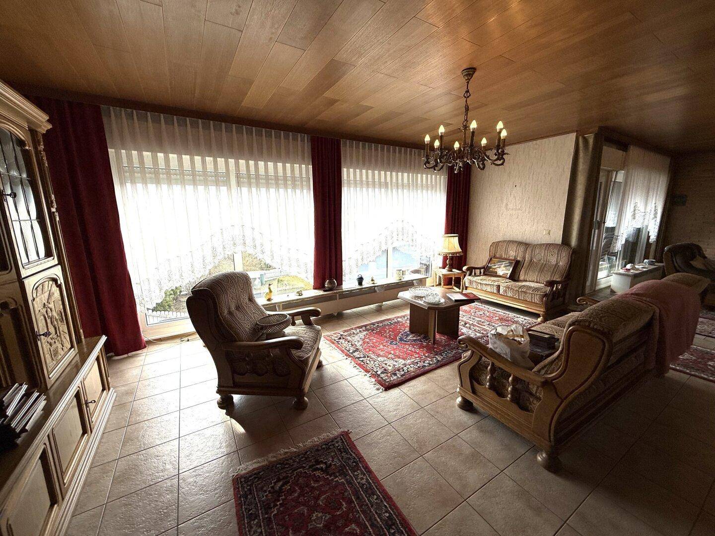 Helles Wohnzimmer mit großen Fenstern, Fliesenboden und gemütlichen Sitzgelegenheiten, lichtdurchflutet.