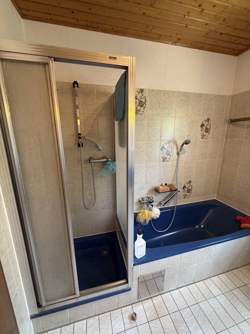 Badezimmer mit Badewanne und Duschkabine, Fliesenboden und Holzdecke, im Stil der 80er Jahre.