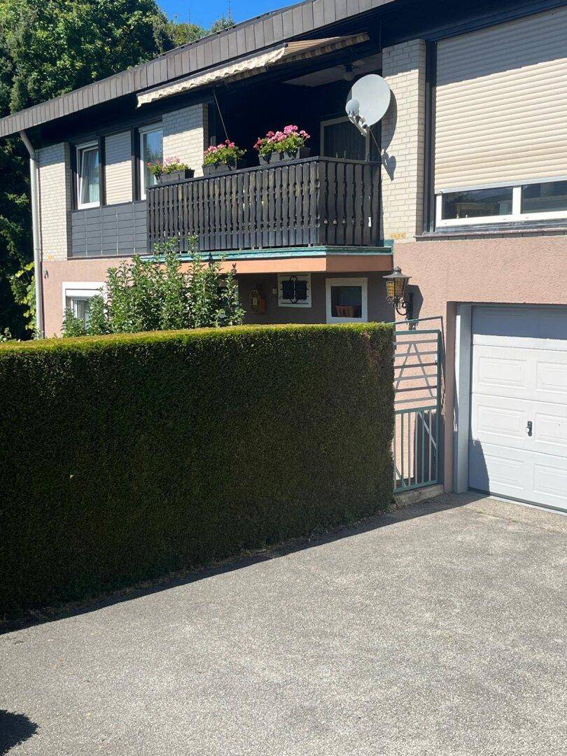 Vorderansicht des Hauses mit Balkon, Hecke und Garagenzufahrt an einem sonnigen Tag.