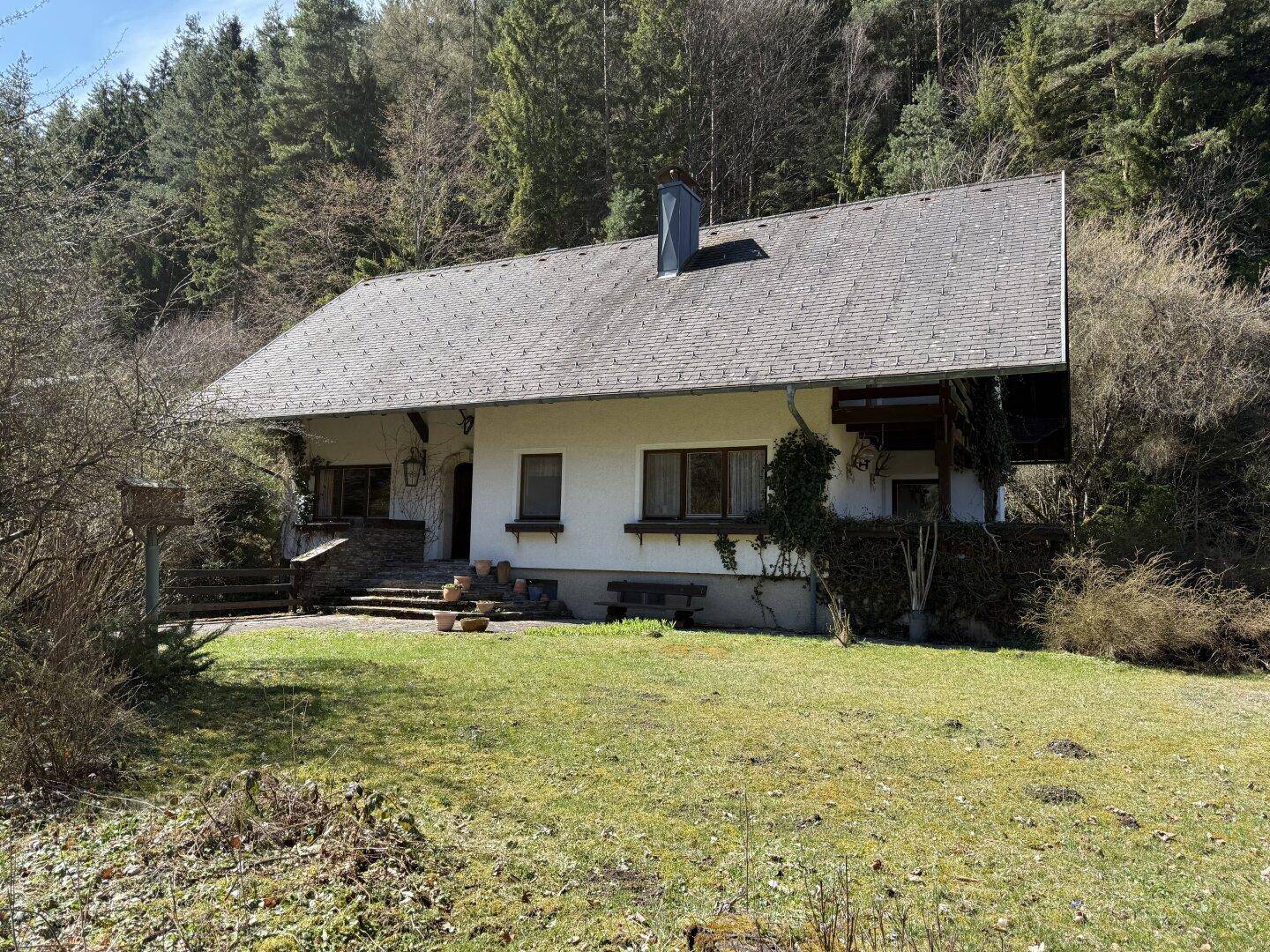 Traditionelles Haus mit weißer Fassade und dunklem Dach, eingebettet in eine grüne, hügelige Landschaft.