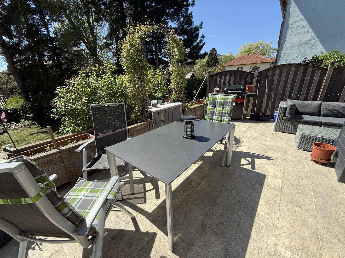 Gemütliche Terrasse mit Esstisch, Stühlen und Lounge-Möbeln, umgeben von Pflanzen und einem Grillbereich.