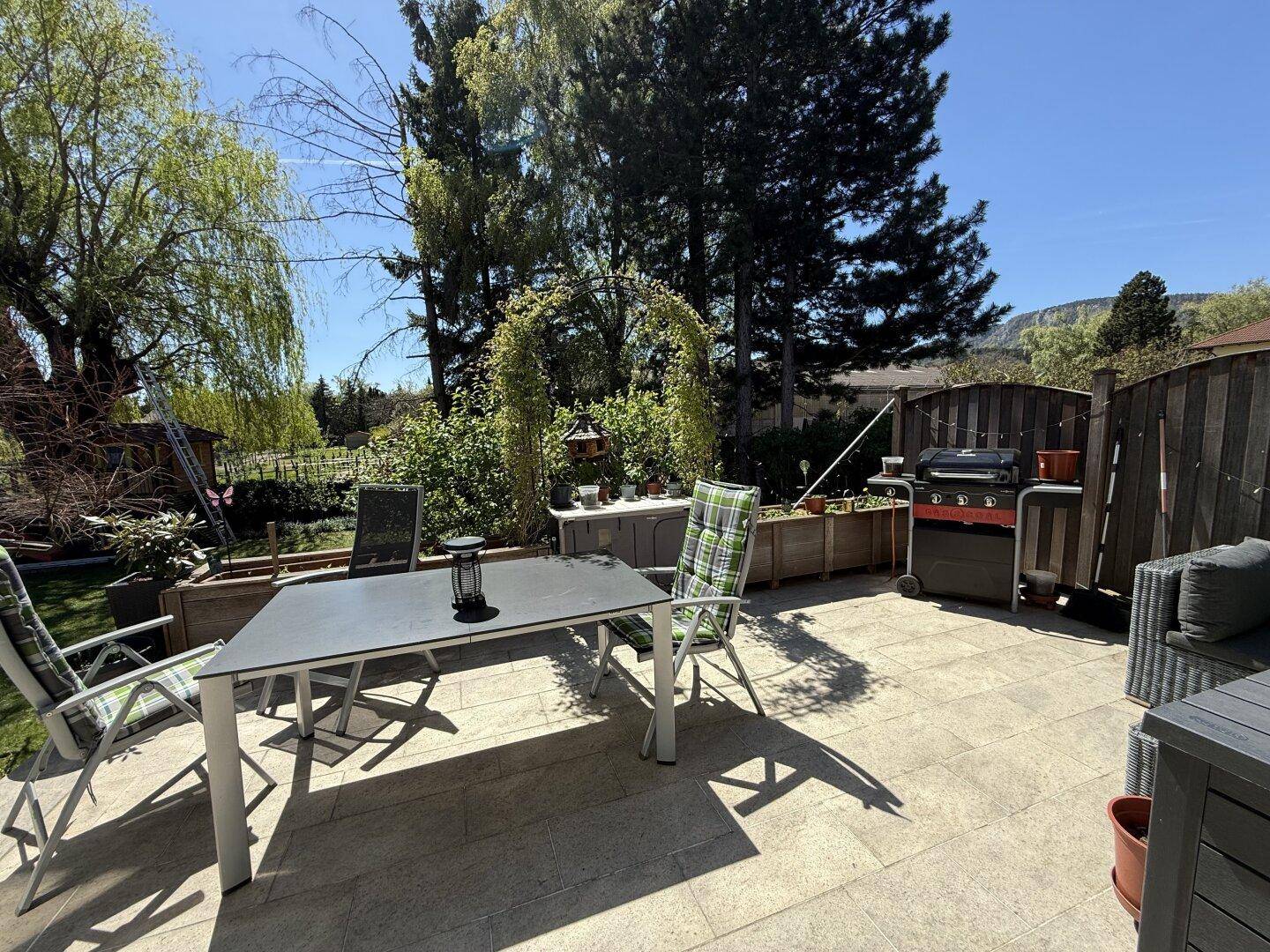 Helle Terrasse mit Sitzgelegenheiten und Grill, umgeben von üppigem Grün und Bäumen.