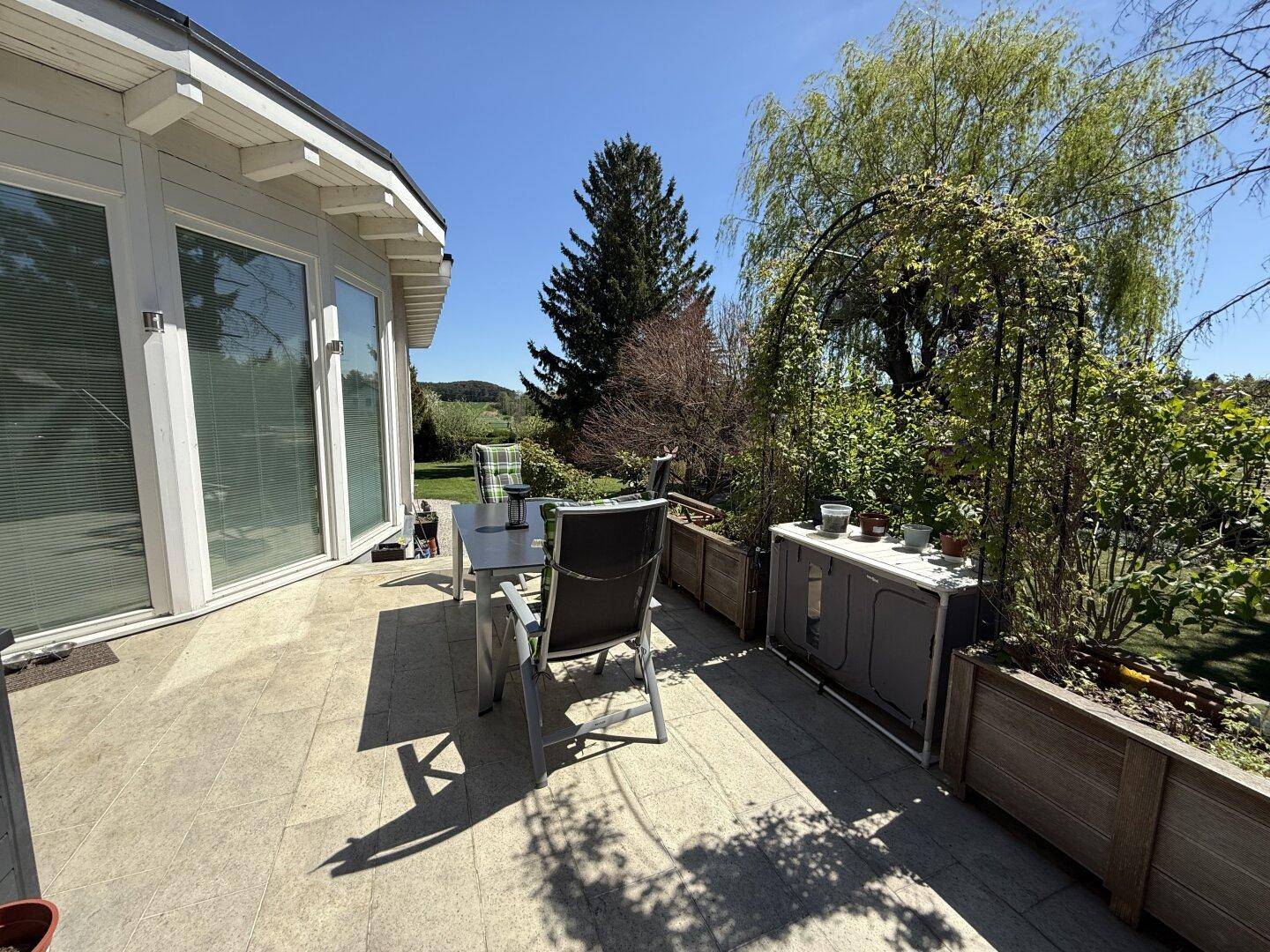 Helle Terrasse mit großen Fenstern zum Haus und weitem Blick über die grüne Umgebung.