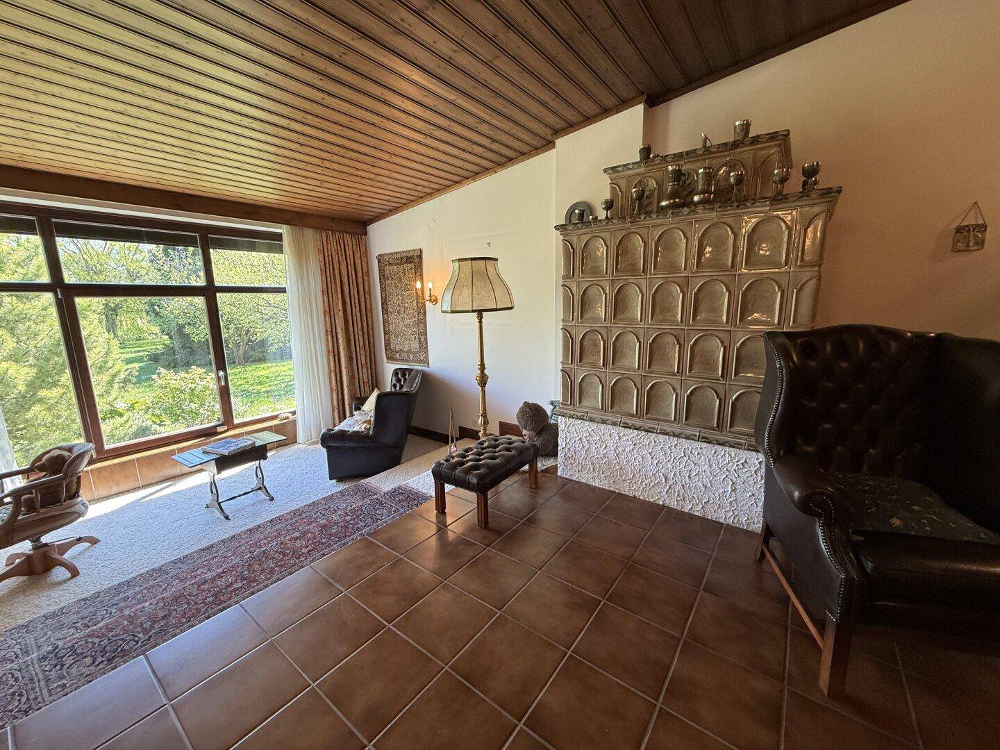 Helles Wohnzimmer mit traditionellem Kachelofen und Blick auf den grünen Außenbereich.