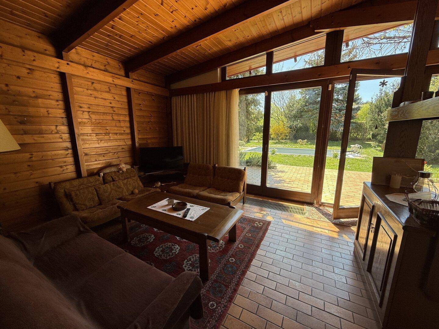 Gemütliches Wohnzimmer mit Holzvertäfelung, großen Fenstern und Blick auf den Poolbereich.