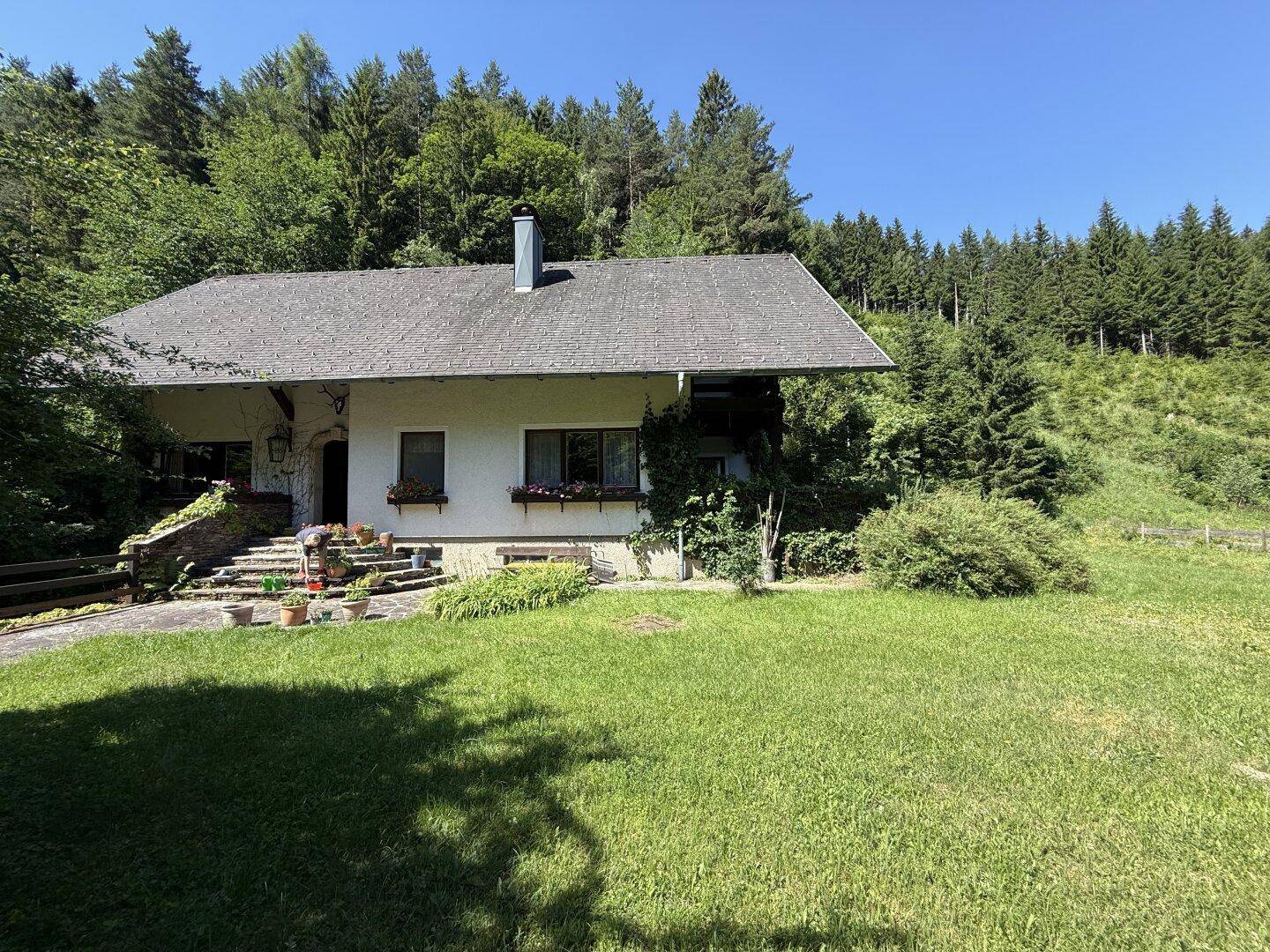 Freistehendes Haus umgeben von weiten Grünflächen und dichtem Wald unter blauem Himmel.