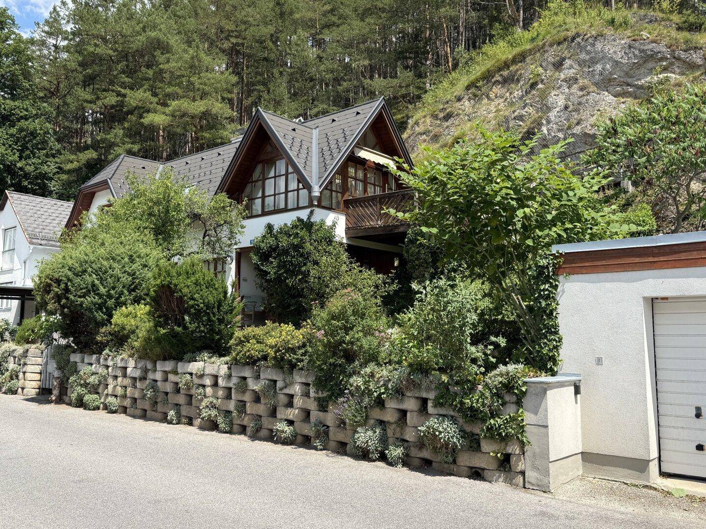Charmantes Haus mit Satteldach und Balkon, umgeben von üppiger Vegetation und Wald.