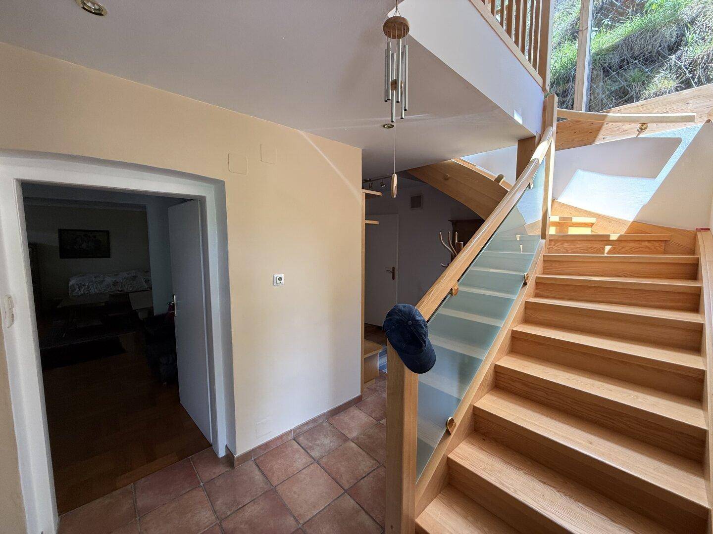 Heller Flur mit Holztreppe, Glasgeländer und Terrakottafliesen im Eingangsbereich.