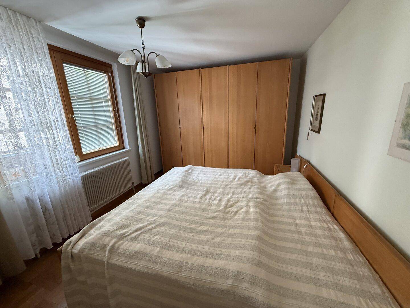 Schlafzimmer mit Doppelbett, Holzschrank und hellem Fenster mit Vorhängen.