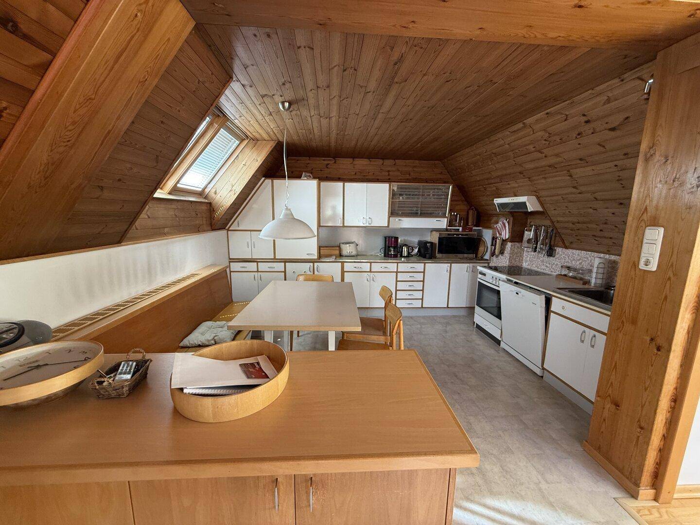 Gemütliche Dachgeschossküche mit Holzbalken, weißen Schränken und Essbereich unter einem Dachfenster.