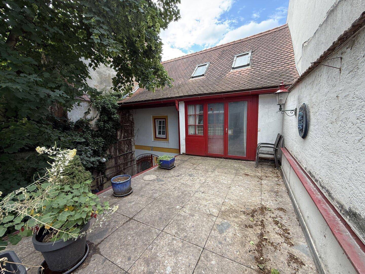 Große gepflasterte Terrasse mit Pflanzen und Zugang zu einem Gebäude mit roter Tür.