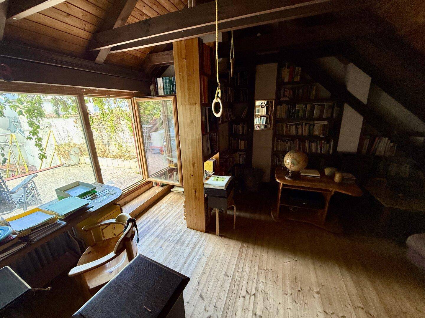Helles Dachzimmer mit Holzbalken, vielen Büchern und großem Fenster zum Außenbereich.