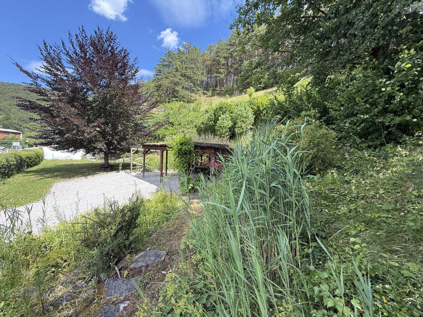 Idyllischer Gartenbereich mit Kiesfläche, Baumbestand und einer kleinen überdachten Sitzgelegenheit.