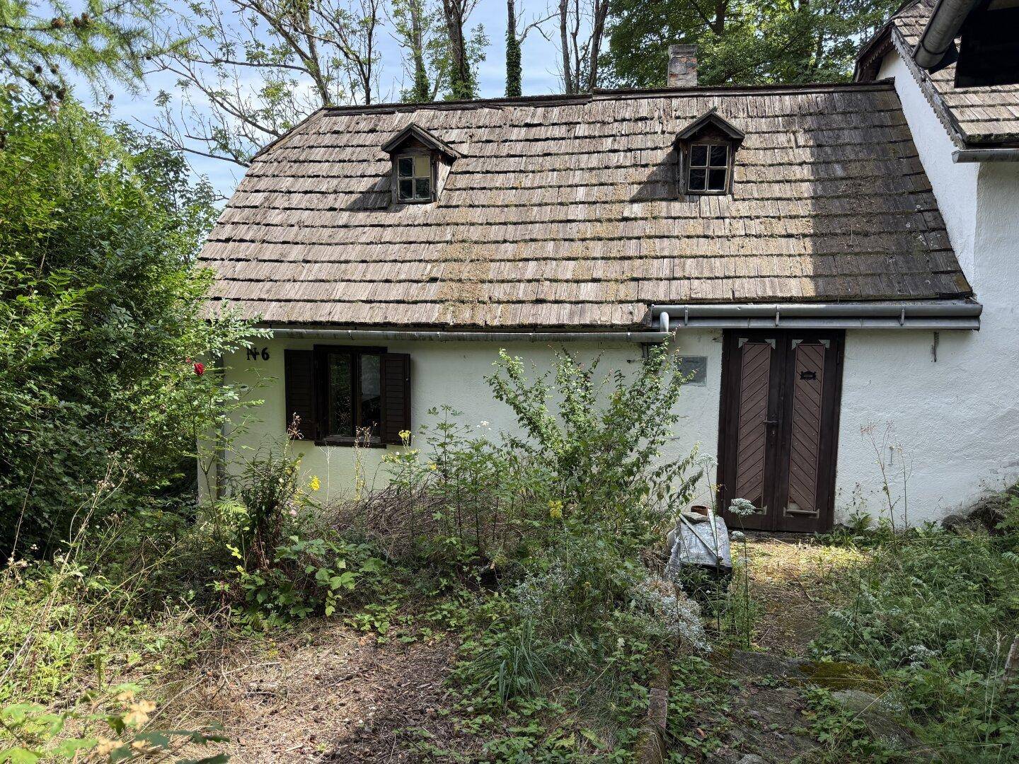 Traditionelles Haus mit weißer Fassade, Holzdach und kleinen Dachgauben, umgeben von Grün.