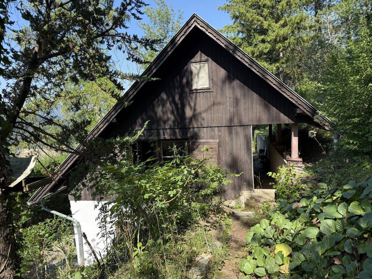 Rustikales Holzhaus mit steilem Giebeldach, umgeben von üppiger Vegetation und hohen Bäumen.