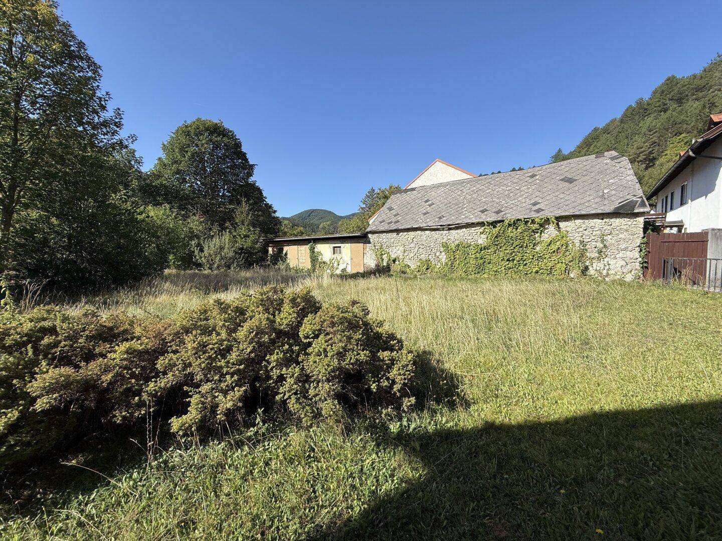 Weitläufige Wiesenfläche mit einem alten Steingebäude und bewaldeten Hügeln im Hintergrund.