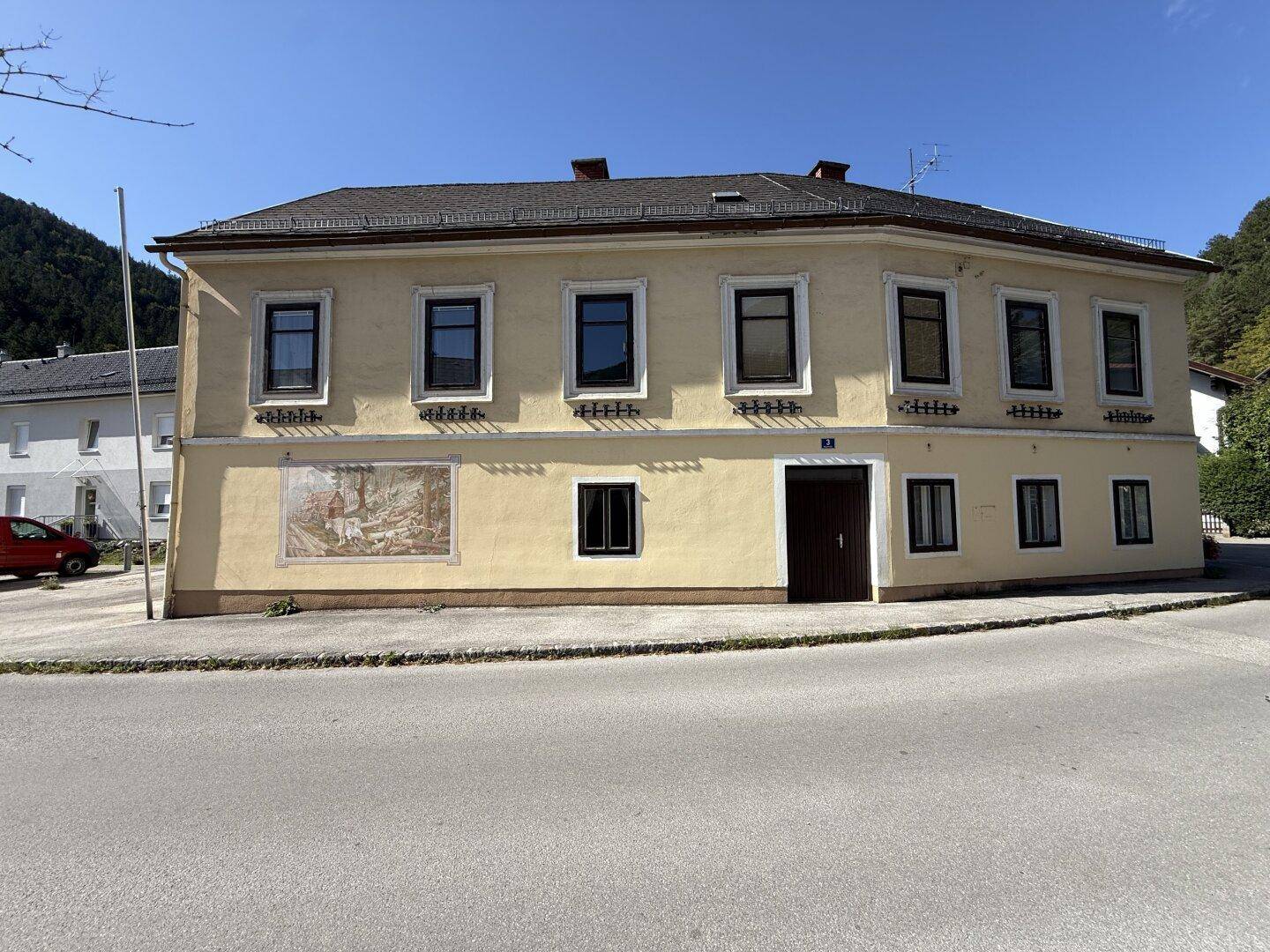 Traditionelles gelbes Gebäude mit dunklen Fenstern und einem Wandgemälde an der Fassade.