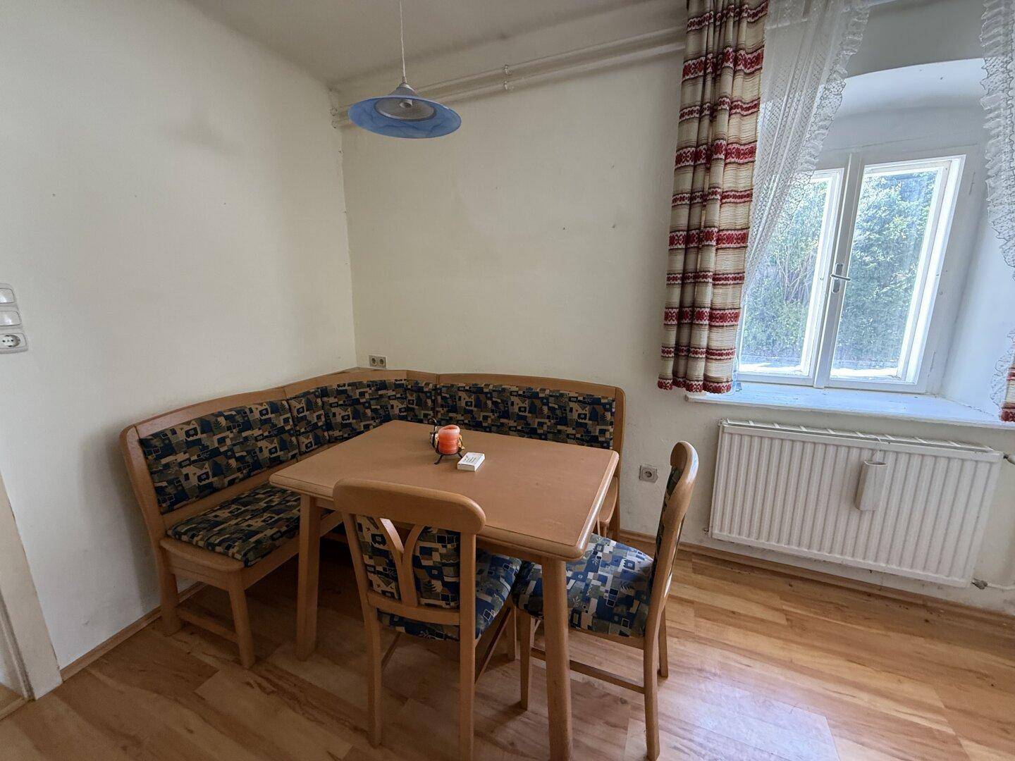 Gemütlicher Essbereich mit Eckbank, Holztisch und Fenster mit karierten Vorhängen.