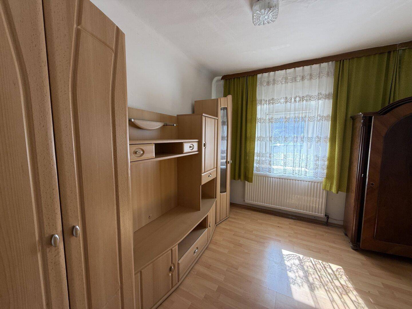 Helles Zimmer mit Holzmöbeln, Laminatboden und einem Fenster mit grünen Vorhängen.