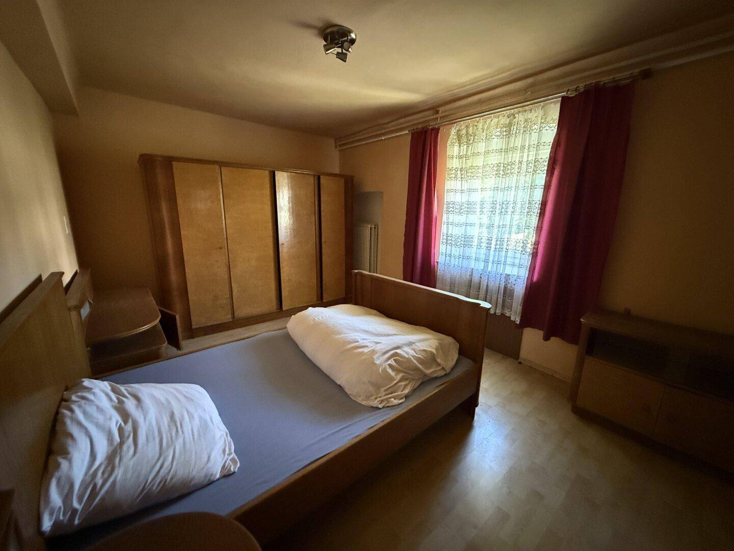 Schlafzimmer mit Einzelbett, Holzschrank und Fenster mit roten Vorhängen.
