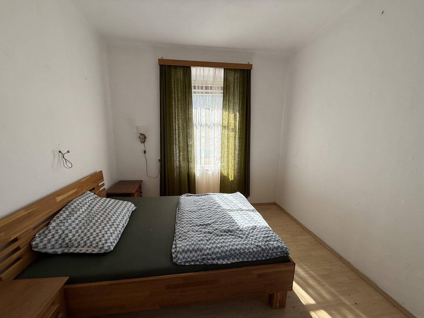 Helles Schlafzimmer mit Holzbett, Nachttisch und Fenster mit dunklen Vorhängen.